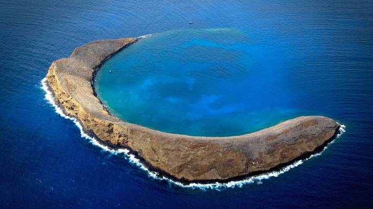 A half moon-shaped crater island surrounded by deep blue water from above