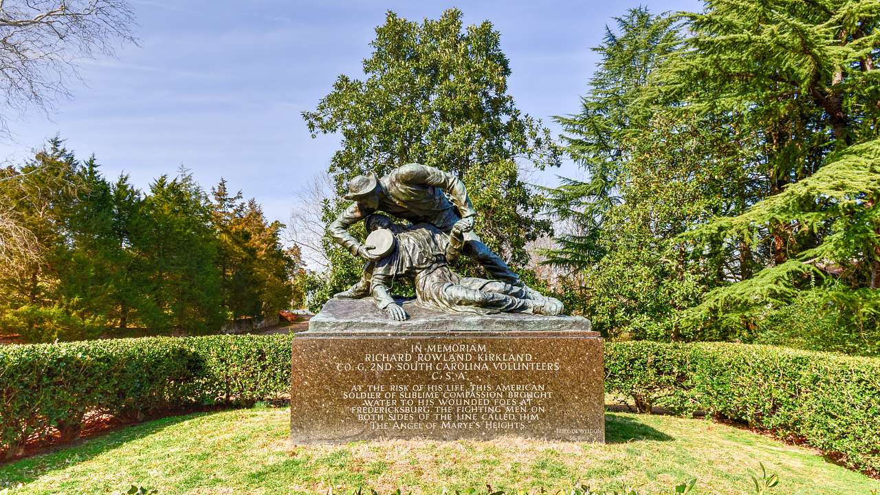 Statue of a soldier holding a man in his arm on a rectangular base, in a garden
