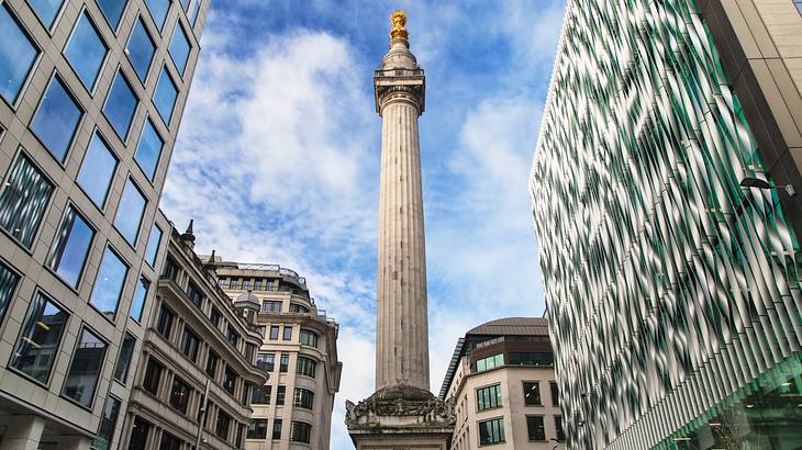 A tall column monument in the middle of buildings under a blue sky with clouds