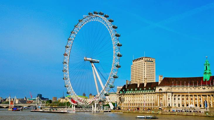 A large Ferris wheel-like structure next to buildings and a river on a clear day