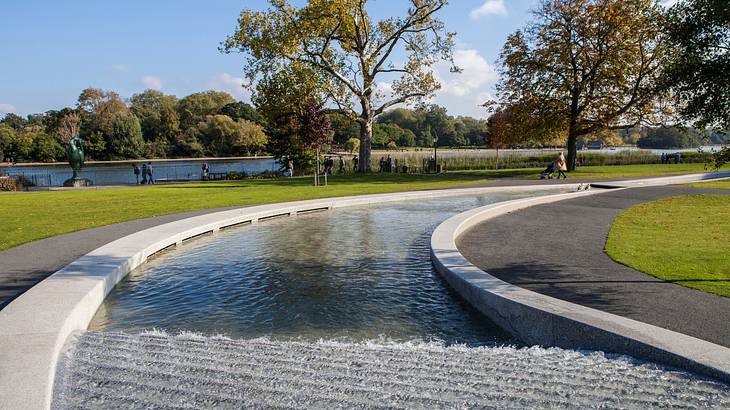 A river-like water fountain next to paths, trees, and green grass