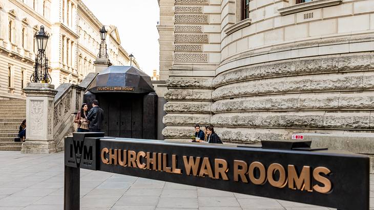 A black and gold sign that says Churchill War Rooms next to stone buildings