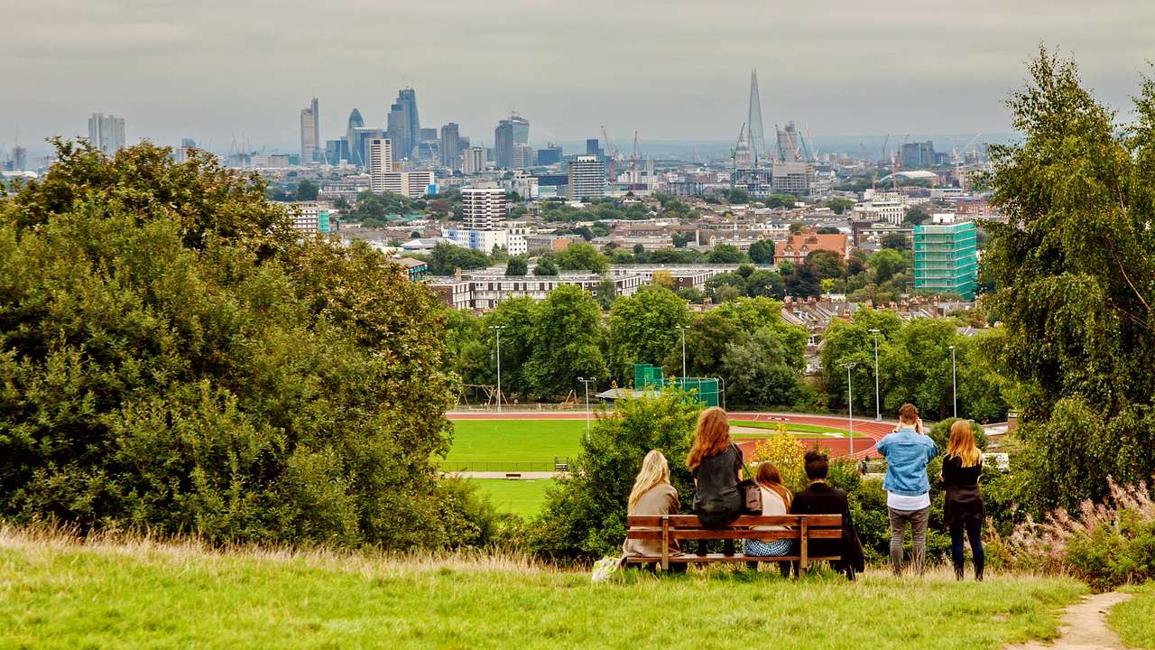 A green hill with trees and people sitting on a bench, and a skyline in the distance