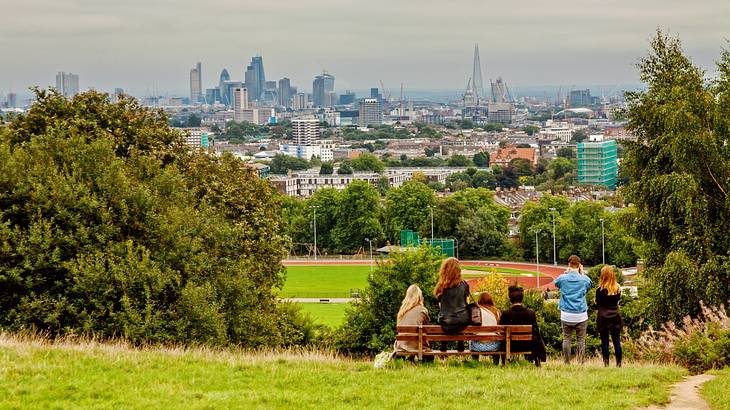 A green hill with trees and people sitting on a bench and a skyline in the distance