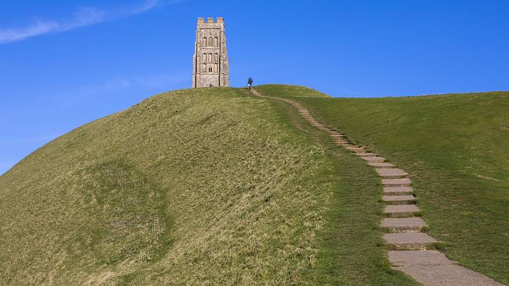 Stairs leading to a topless, stone tower atop a grassy hill under clear blue skies