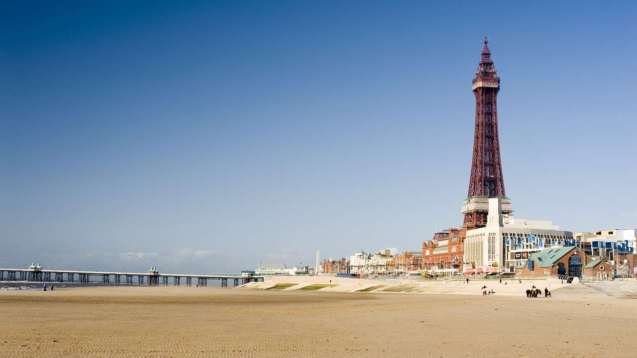 A tall red pointy tower surrounded by buildings overlooking a sandy beach with a pier