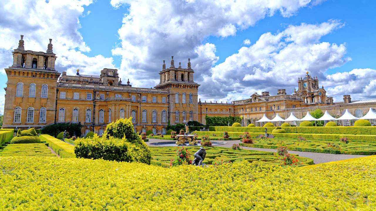 A large castle with a lush garden containing yellow hedges in front on a sunny day