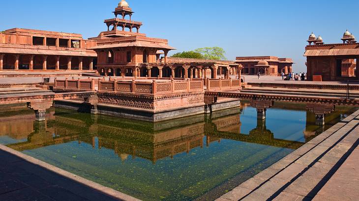 A complex of red sandstone buildings with a pool of water in front on a sunny day