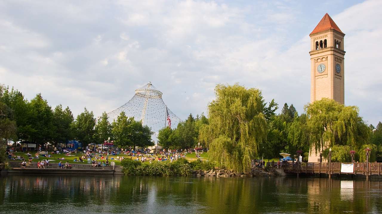 A lake next to grass with people on it, trees, and a tower with a clock