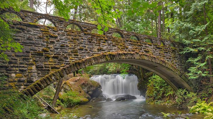 A stone bridge over a river and a small waterfall surrounded by trees