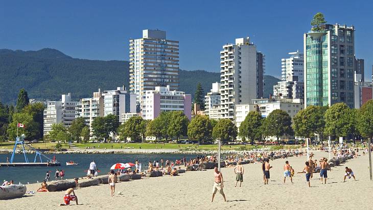 A beach with people playing volleyball next to the ocean, buildings, and mountains