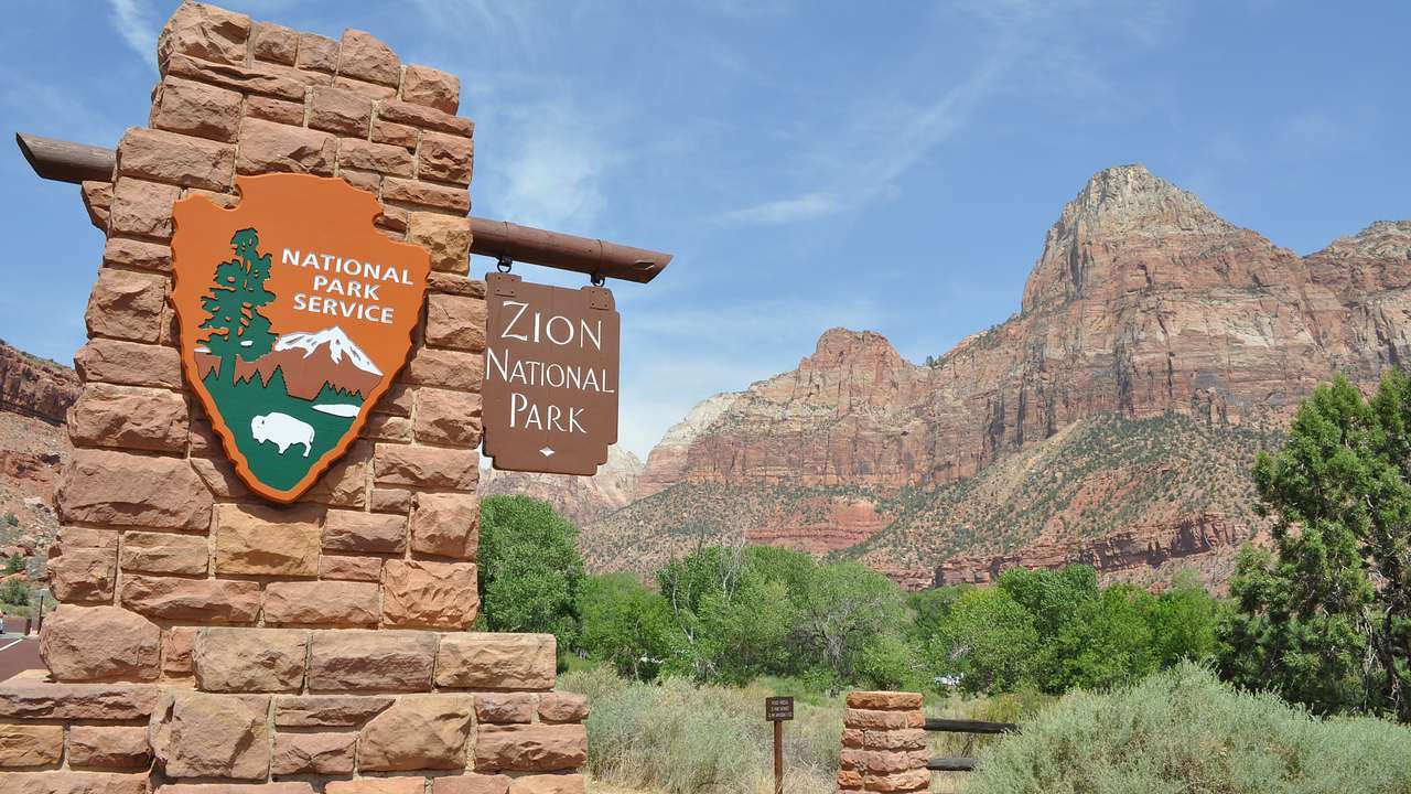 A sign "Zion National Park" on a rock wall with trees and mountains in the background