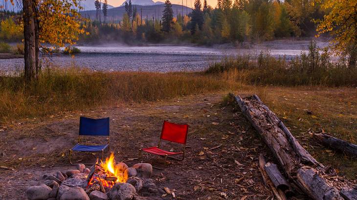 A fire pit with camping chairs and a lake with trees and mountains in the distance