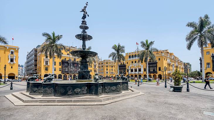 A fountain in a plaza surrounded by yellow buildings and palm trees on a sunny day A fountain in a plaza surrounded by yellow buildings and palm trees on a sunny day