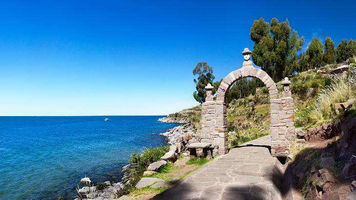 A stone archway on a hilly path with trees looking out onto a blue body of water