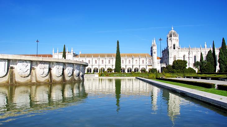 A white monastery structure next to trees, greenery, and a lake on a clear day