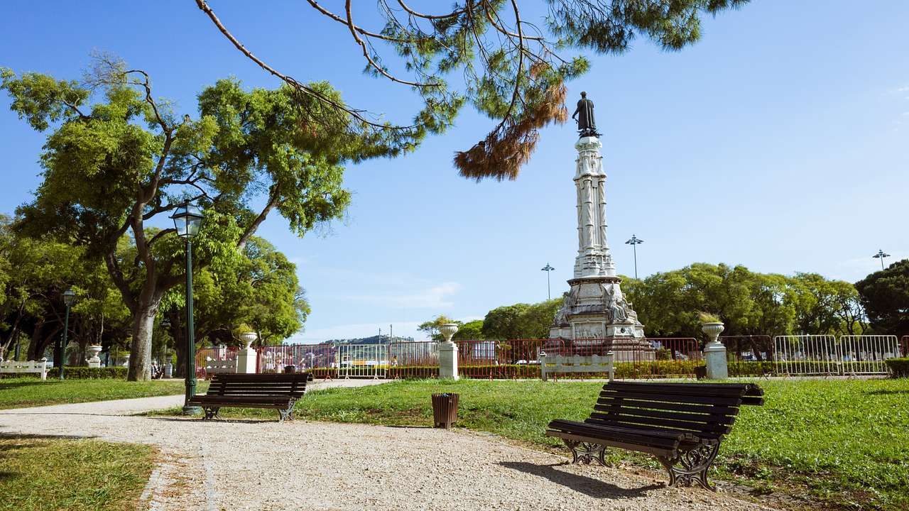 A park with a gravel path, greenery, benches, and a statue on an obelisk