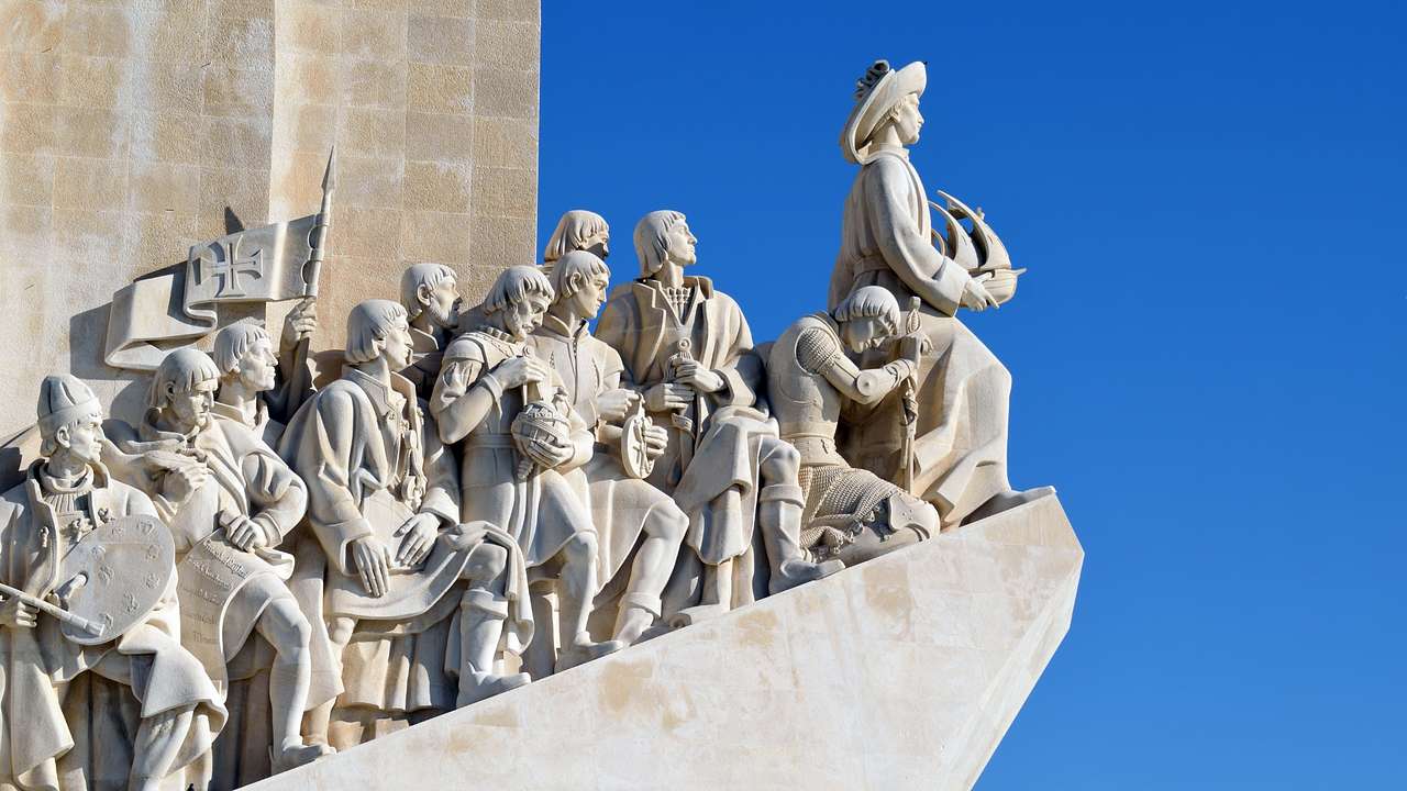 A large statue with men holding flags and ships next to a wall and a clear blue sky
