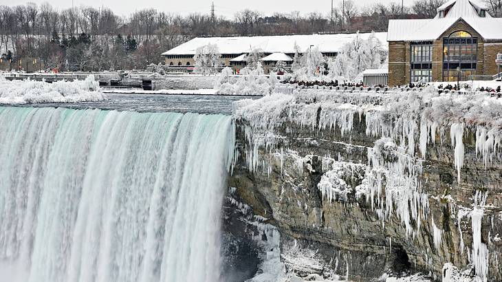 A large waterfall with snow on the sides and buildings in the background