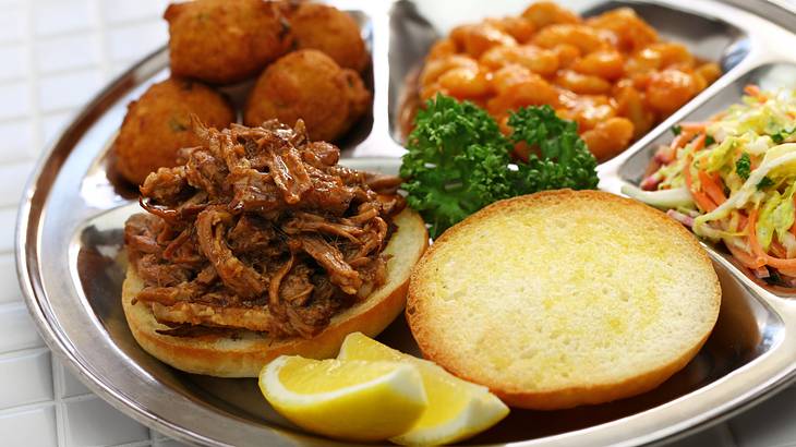 Pulled pork, hush puppies, baked beans, coleslaw, lemon slices, and bread on a plate