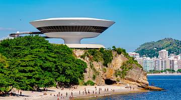 A white spaceship-style building on a green hill overlooking blue water and sand