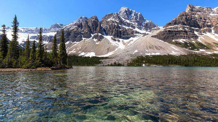 Snow-capped mountains and rocky cliffs with trees at the base facing a lake