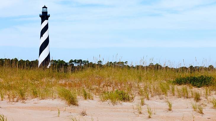 A black and white striped lighthouse on a sandy ground with grass and greenery behind