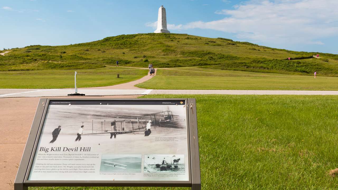 A sign with a picture of a monument, and a road going towards a hill with a pillar