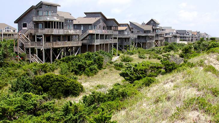A row of beach houses surrounded by green bushes and grass facing a low hill incline