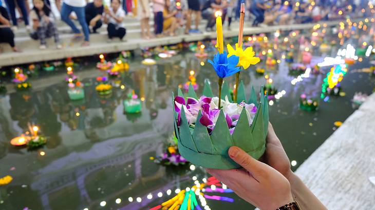 A hand holding a candle with flower petals in front of canal full of floating objects