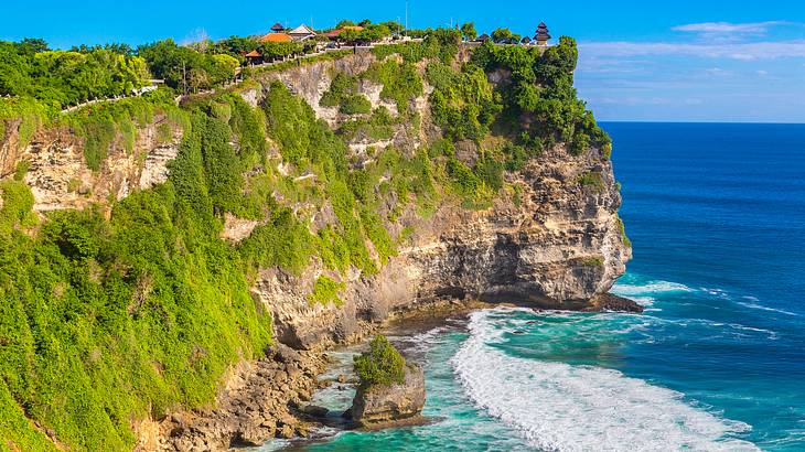 A steep cliff's edge with grass, small temples on top and waves below