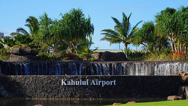A stone wall sign "Kahului Airport" with trees and a fountain behind it