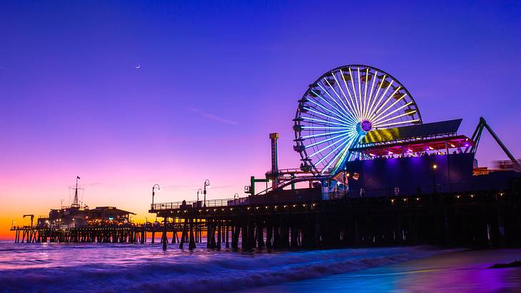 A pier with lapping waves and a lit up Ferris Wheel surrounded by a purple sky
