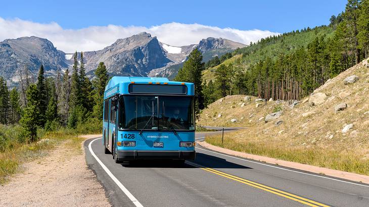 A blue shuttle bus driving down the road with trees and mountains in the background