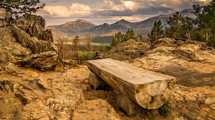 A wood bench on a rocky hill with trees and mountains in the distance