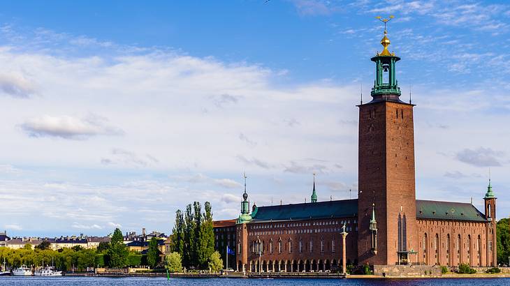 A red brick building with a tower next to green trees and the water
