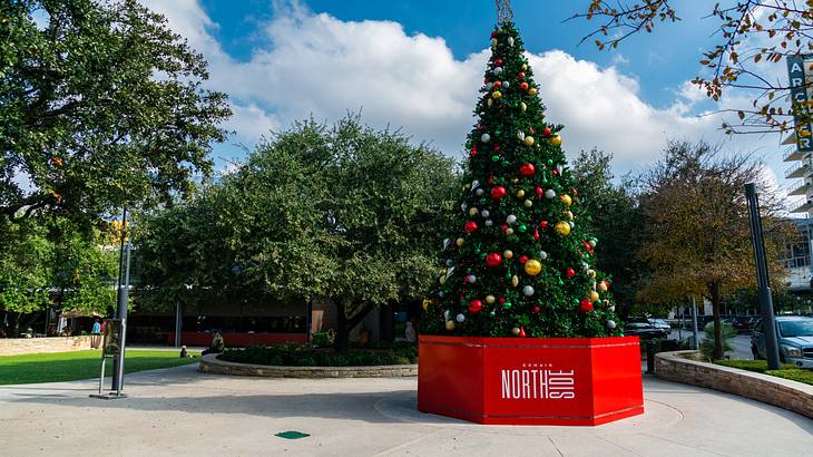 A large Christmas tree with decorations on a red stand with trees in the background