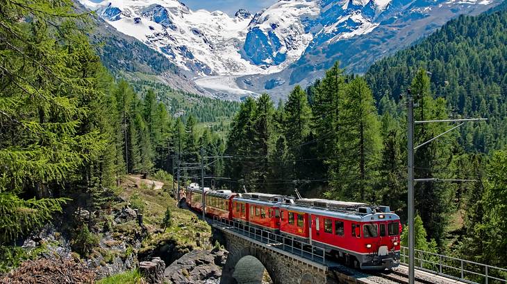 A red train on a track through alpine trees next to a snow-covered mountain
