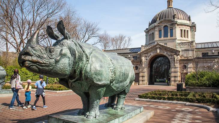 A statue of a rhino in front of an old building with three people walking towards it A statue of a rhino in front of an old building with three people walking towards it