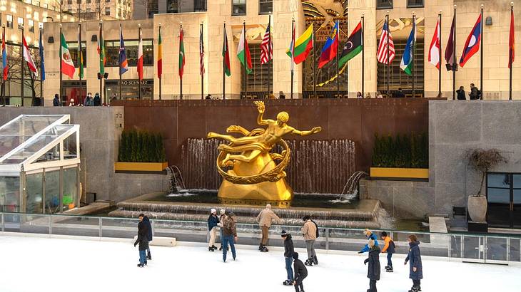 An ice rink with people skating with a statue, flags, and buildings behind it An ice rink with people skating with a statue, flags, and buildings behind it