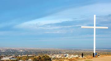 A hilltop with a large white cross on it and views across a town under a blue sky