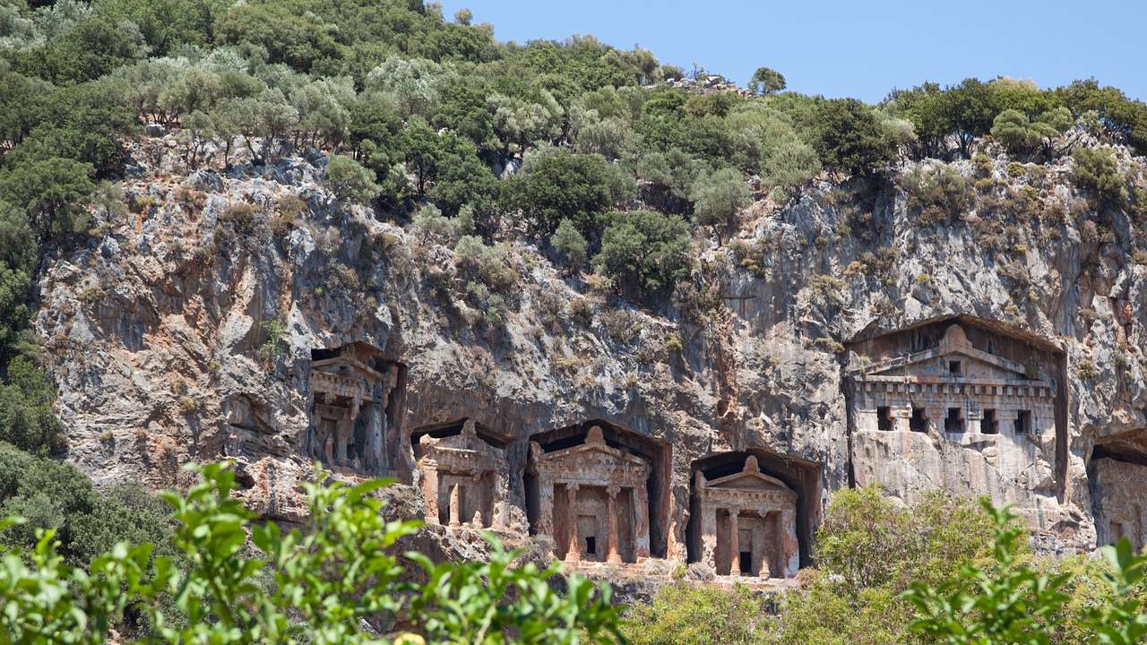 Tombs with columns carved into a mountains with trees around it