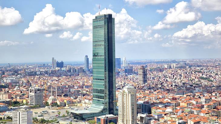 A tall skyscraper surrounded by smaller city buildings under a blue sky with clouds