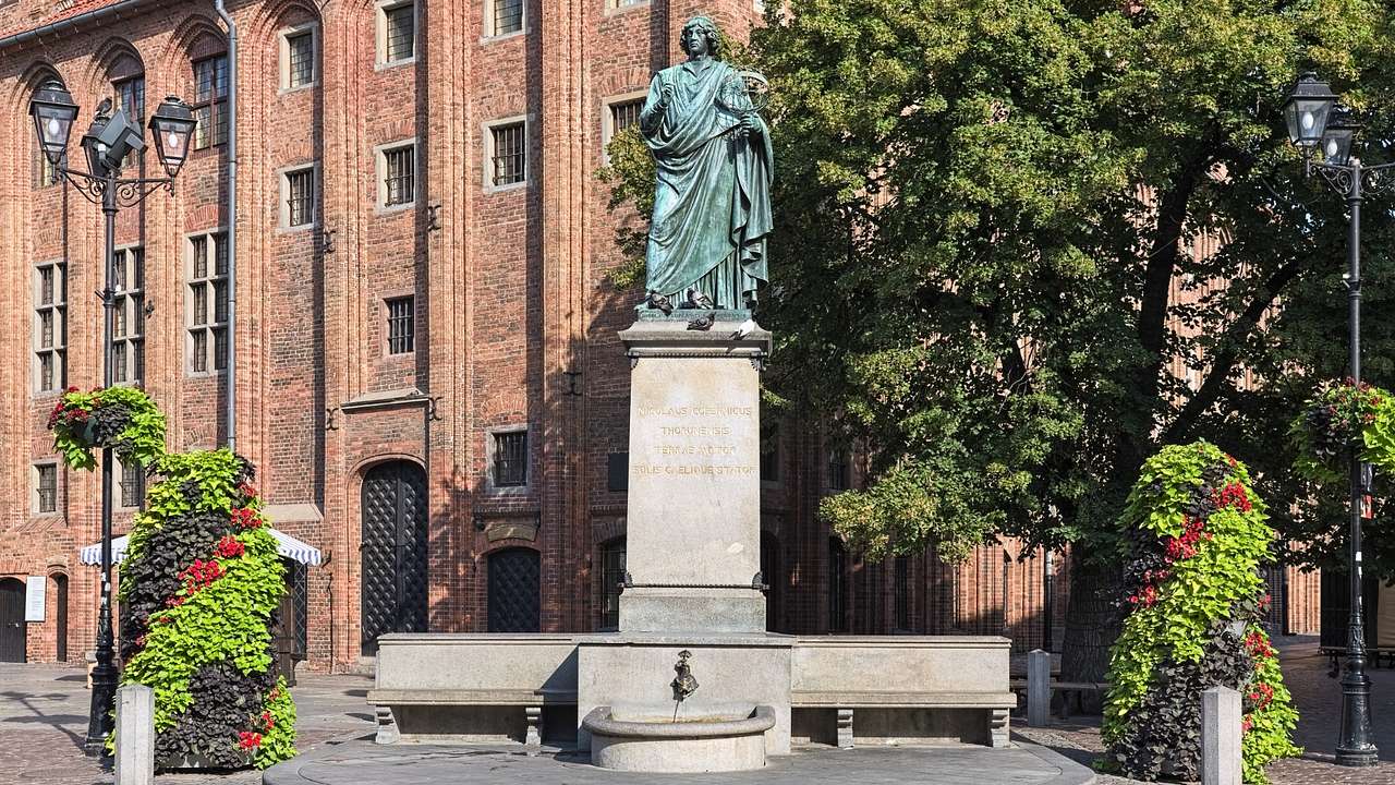 An oxidized statue of a person on a stone pedestal next to a red brick building