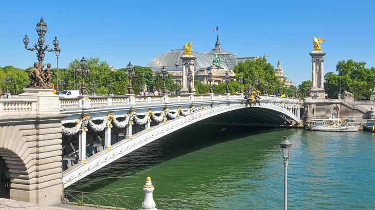 An arched bridge with lamp posts atop a flowing river with two gold sculptures