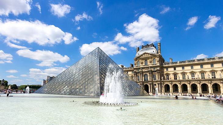 A domed building facing a glass pyramid and a fountain under sunny skies