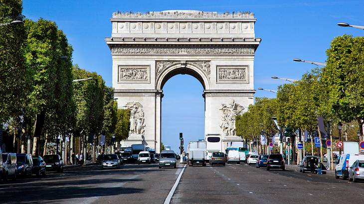 A white, arched stone monument with carvings facing a road with cars lined with trees