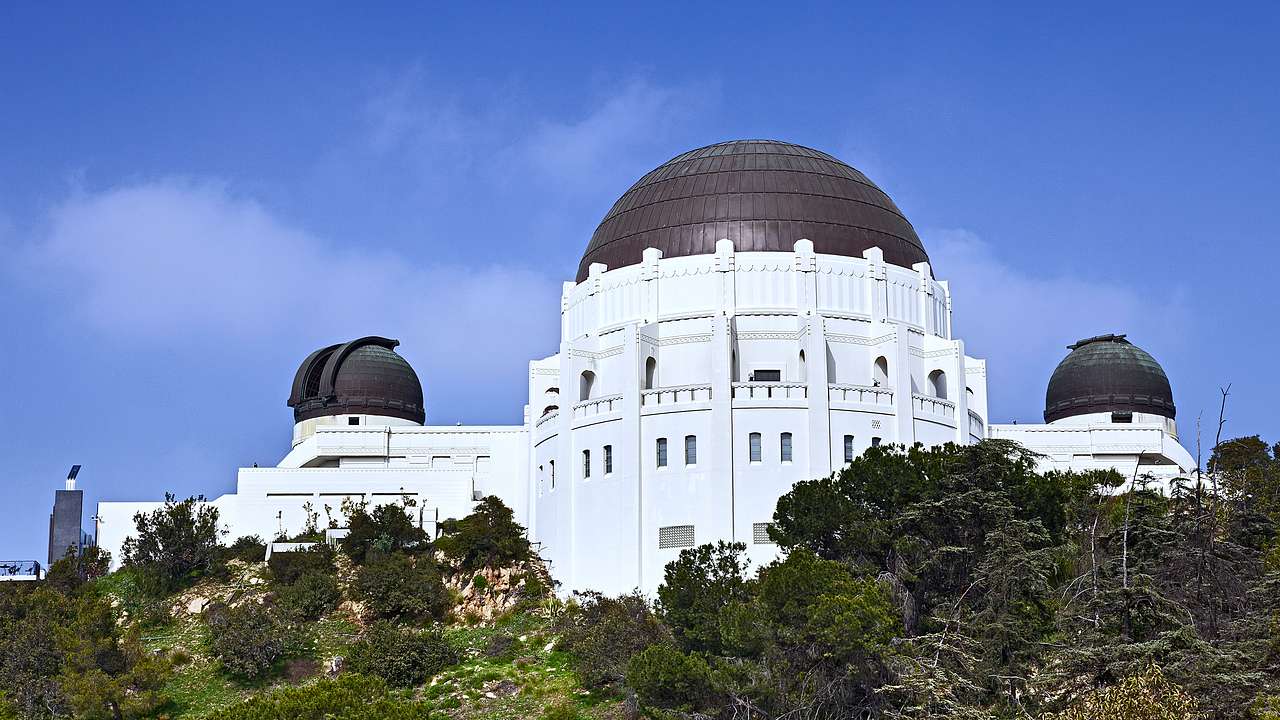 A white building with three black domes on top of a tree-covered mountain