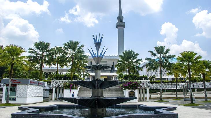 View of a mosque's entrance from its stairs with palm trees and a fountain in front