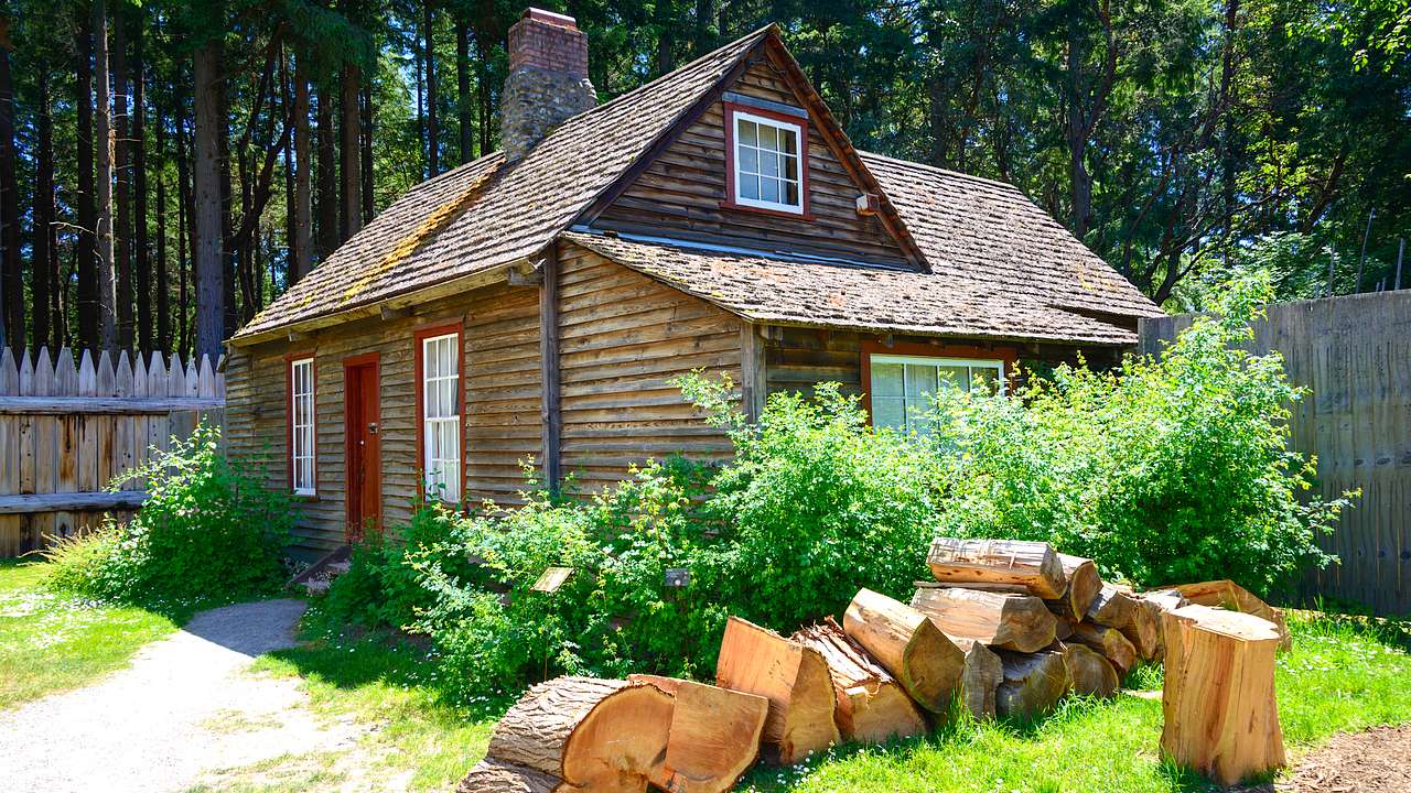 A log cabin with a wooden fence at the back and a pile of large logs in front of it